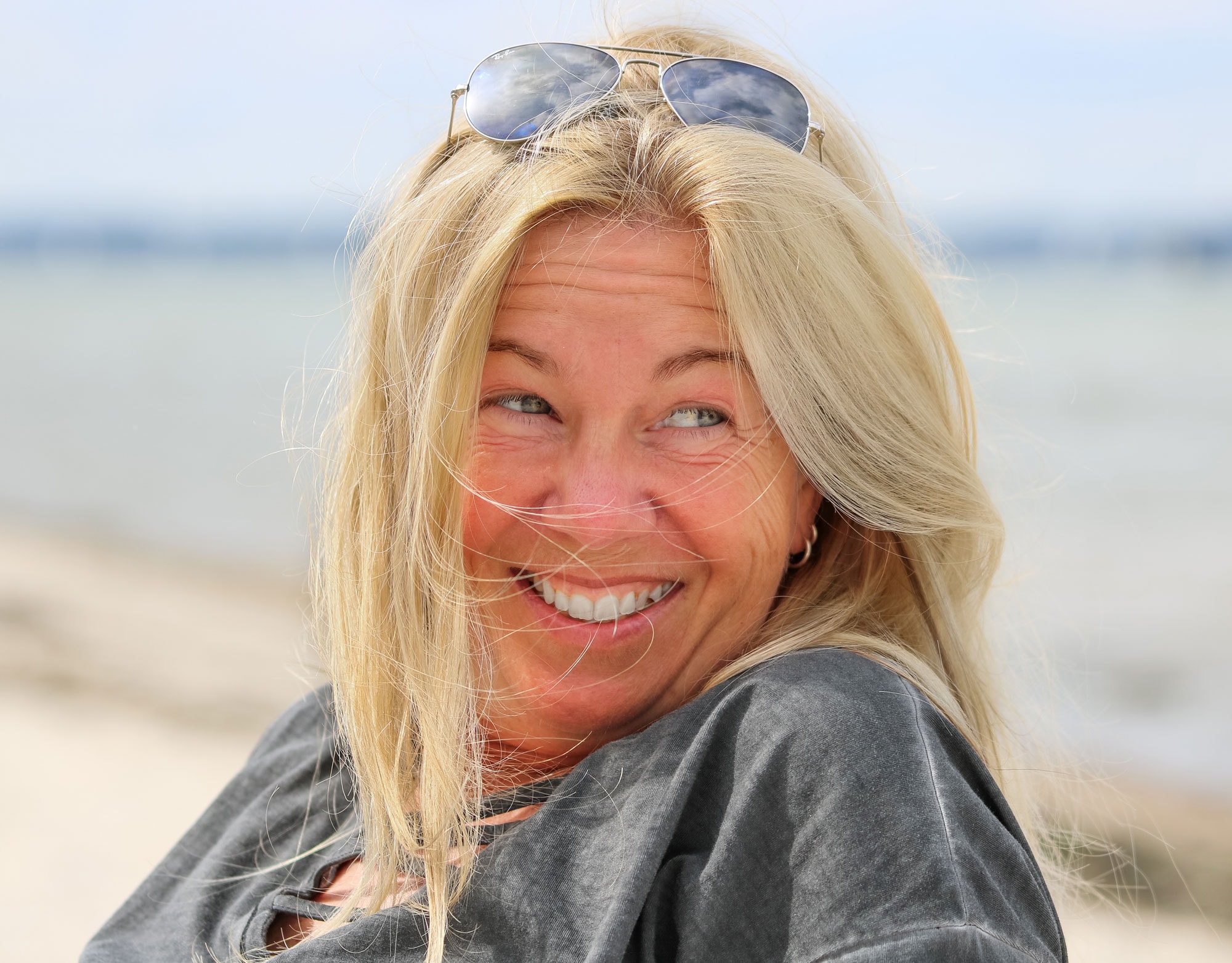 Hochzeitsfotografin Fürstenfeldbruck Hochzeitsfotografin Diane K. Senft im Selbstporträt. Mit Sonnenbrille im Haar am Strand, lachend und entspannt.