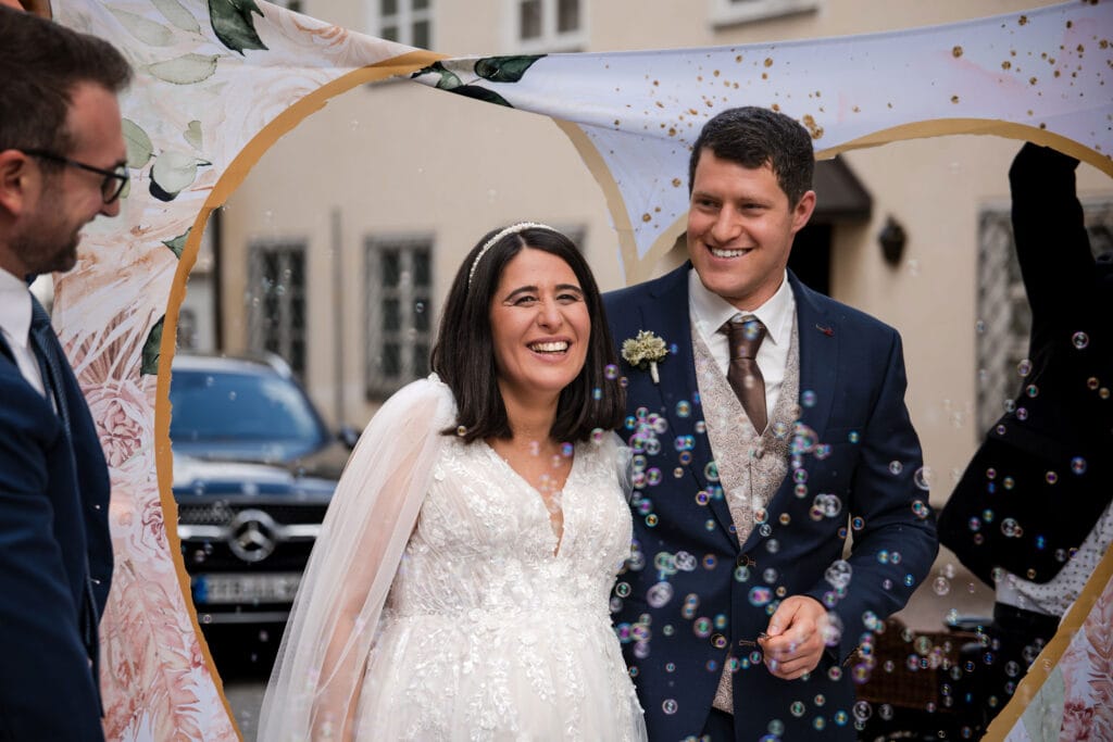 Hochzeitspaar mit lachendem Bräutigam und Braut unter Blumenbogen, festliche Stimmung, outdoor.