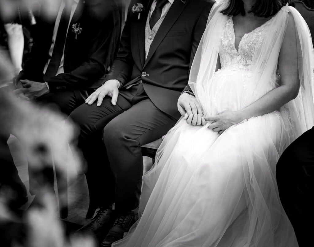 Braut in weißem Kleid bei Hochzeit, sitzend mit Gästen, schwarz-weiß.