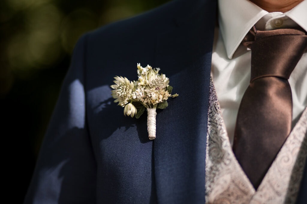 Nahaufnahme eines Bräutigams in dunkler Anzugjacke mit Blumenbrosche an der Schulter.